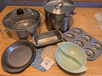 Overview of two large cooking pots with lids, various bakeware including muffin tin, bread tin, pie tin, and a divided serving bowl on a wooden table.