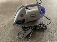 White and blue Bissell Spot Lifter carpet spot cleaner device lying on carpet with power cord coiled beside it.