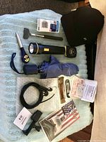 Photo showing the full lot laid out on light blue fabric background including black cap, compact umbrella, flashlight, measuring tape, back scratcher, tie down strap, penknife, screwdriver, first aid kit, tissue pack, NJ map, and cell phone holder