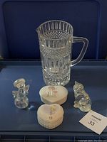 Full lot arranged on blue background showing crystal pitcher, two Shannon Crystal figurines, two Precious Moments heart trinket boxes, and lot number tag.