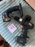 Photo showing Black & Decker 6V cordless drill with charger and Ryobi flashlight resting on fabric surface.