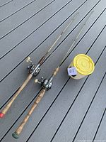 Photo showing two vintage fishing poles with cork handles and baitcasting reels, next to plastic minnow bucket with yellow lid on wood deck.