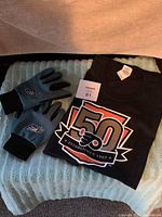 Photo of the folded black Flyers 50th Anniversary T-shirt and a pair of blue and black XLEAGLES work gloves laid on fabric.