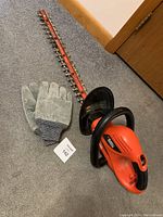 Full view of electric hedge trimmer and protective gloves on carpeted floor, showing blades and handle details.