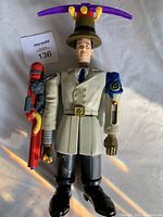Full front view of Inspector Gadget figure showing beige trench coat, black boots, red grappling hook arm attachment, and blue mechanical glove on other arm, with rotor helicopter on brown hat.