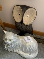 Photo showing the large dark brown owl wall hanging with big cream eyes alongside the white glossy cat lawn ornament lying down.