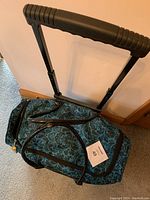 Top view of teal and black patterned carry-on bag with extended black telescoping handle and two black fabric carrying straps.
