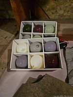 Photo of 9 assorted colored glass ornaments in white box compartments showing colors: green, purple, light green, white, gray, lavender.