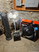 Photo of all items in the lot on a basement floor against brick wall: Seville tower fan, white standing fan, Master Craft heater, and boxed Honeywell Turbo Force fan.