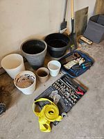 Photo showing garden pots, gloves, ratchet tool set, shovel, and yellow ratchet strap all arranged on floor in garage setting.
