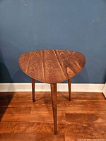 Front view of the walnut side table showing the triangular top and three legs on wood flooring against blue wall.