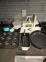 Kitchen gadgets and bakeware including mandolin slicer, spiralizer, muffin tin, cake pans, whisk, measuring cups, and other tools arranged on kitchen counter.