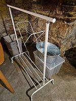 White metal clothes hanging rail, drying rack, laundry basket, metal bucket inside a plastic bin in a basement with stone wall background