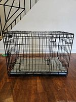 Front view of black metal MidWest iCrate dog crate with grey dog bed visible inside on wooden floor under stairs.