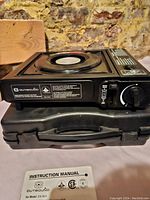 Side view of the black single burner gas stove sitting on top of a black plastic carrying case with instruction manual in front.