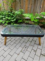 Rectangular outdoor coffee table with black ribbed base and four wooden legs on a stone patio, partially surrounded by plants.