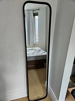 Full view of the full-length leaning mirror placed against a white wall next to a bed with a window reflecting in the mirror.