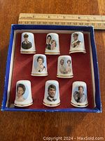 Top down view of the 9 bone china thimbles with portraits of British royals arranged in a box with red felt lining and a blue border around the edge.