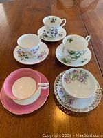 Five china cups and saucers displayed on a wooden table, showing floral decorations and color variations including pink and purple.