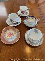 Five china cups and matching saucers displayed on wooden table showing different floral designs and gold trim.