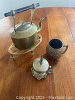 Photo showing vintage brass teapot with black handle on a brass stand, a brass mug with pattern, and a decorative bell with detailed base.
