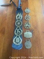 Overall view of 9 vintage horse brasses and leather mounting belt on wood surface.