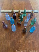 Top-down view of 16 assorted mini glass bottles arranged on a wooden surface with ruler showing size approximately 2 inches tall. Bottles are in amber, blue, green, and clear colors, most have cork stoppers.