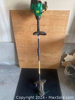 Full view of the Weed Eater string trimmer showing its green and black body, attached cutting guard and shaft, supported upright against a plywood board.
