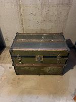 Closed view of green metal antique trunk showing front positioned on concrete floor against wall.