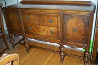 Front angle view of 1940's walnut buffet showing detailed wood carvings on side doors, two center drawers with round metal knobs, turned legs and connecting stabilizing bars.