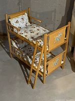 Stacked wooden bunk beds for dolls with attached ladder and mattress with patterned bedding.