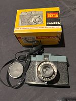 Top view of Diana camera, lens cap, and original yellow box with branding and camera image.