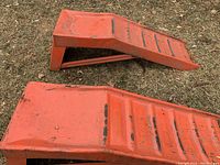 Two red metal car ramps laying on grass, showing stepped incline surface with visible scratches and paint wear.