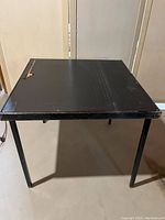 Front view of the square black folding card table showing worn top and scuffed edges