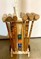 Front view of white wooden croquet cart holding 8 wooden mallets and 6 croquet balls underneath.