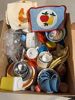 Top-down view of assorted doll dishes and accessories inside a box, showing various plastic and metal items including cups, plates, spoon, trays, and cloths.