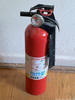 Photo of a red Kidde fire extinguisher with a black handle and safety pin against a white textured wall.