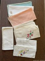 Photo showing 8 assorted linen handkerchiefs in different colors (green, white, pink, cream, off-white), several with detailed embroidery including floral and monogram motifs, some with lace edges.