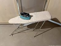 Children's play iron and ironing board set shown side-by-side on floor against wall. Ironing board has white cloth cover and metal frame.