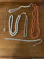Photo showing six vintage jewelry pieces arranged on a wooden surface: coral bead necklace, two white bead necklaces, clear bead necklace, and three silver-tone chain items including two bracelets and one chain with pendant.