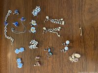 Photo shows nine sets of vintage clip-on earrings and one pin/brooch on a wooden surface. Includes blue heart earrings, floral designs, pearl dangle earrings, a silver bracelet, and a small leaf shaped pin.