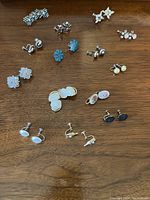 Top-down photo showing 14 pairs of vintage clip-on earrings arranged on a wooden surface including floral and stone styles.