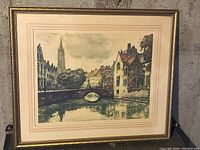 Framed artwork showing a street scene with castle-like home and canal with bridge, 25 x 22 inches.
