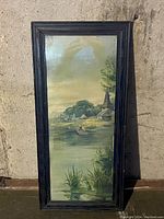 Framed picture depicting a boy sailing a small sailboat on a lake with houses, greenery, and a church steeple in background.