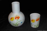 Photo showing enamel flowered pitcher and drinking glass front side with colorful red, orange, yellow, and blue floral motifs on white background.