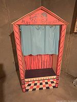 Front view of the puppet show theater showing the painted cardboard frame and blue plastic curtain with theatrical masks and 'Play Theater' text.