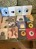 Top-down image showing 16 individual 45 RPM vinyl records in various original paper sleeves and covers arranged on a patterned couch.