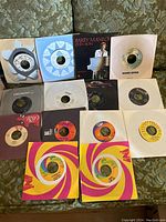 Top view of 14 vinyl 45 RPM records laid out over fabric surface showing record labels and sleeves