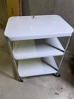 Front angled view of white rolling table with 2 shelves showing the top, shelves, and metal legs with caster wheels.