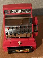 Photo of red toy cash register made of metal and plastic, showing numbered buttons, price display, small white knob, and pull-out drawer with colorful graphic.